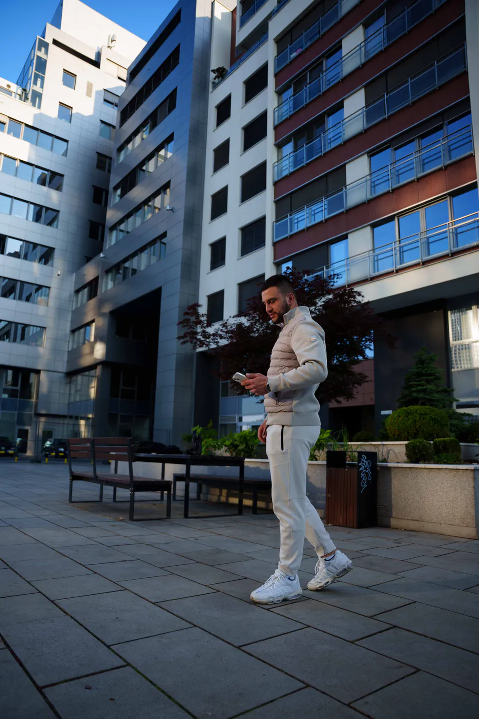 Man in a cream tracksuit walking across a paved plaza at dusk, glass and red-panelled office blocks behind him