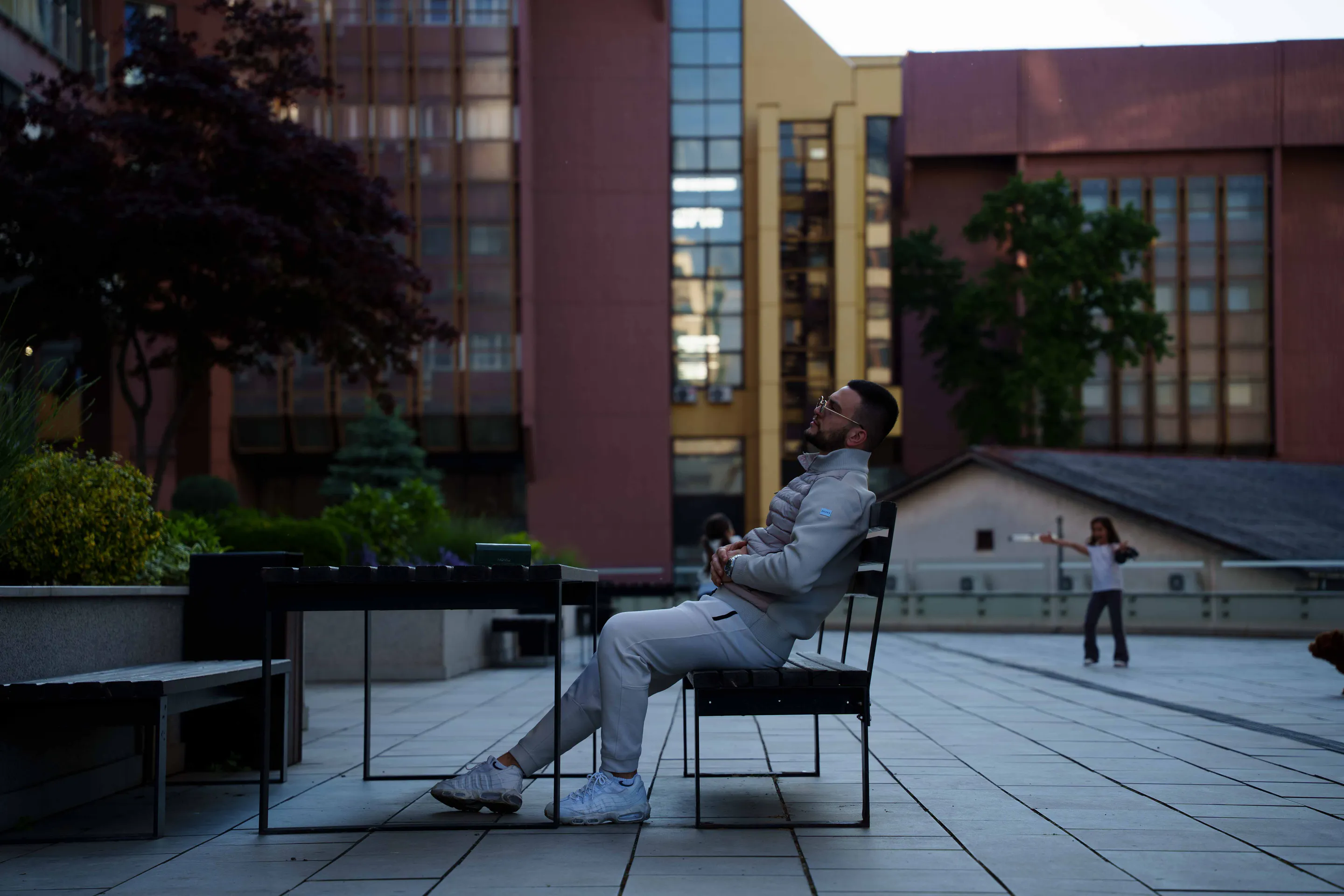 The plaza at dusk — reclined on the bench, small against the buildings