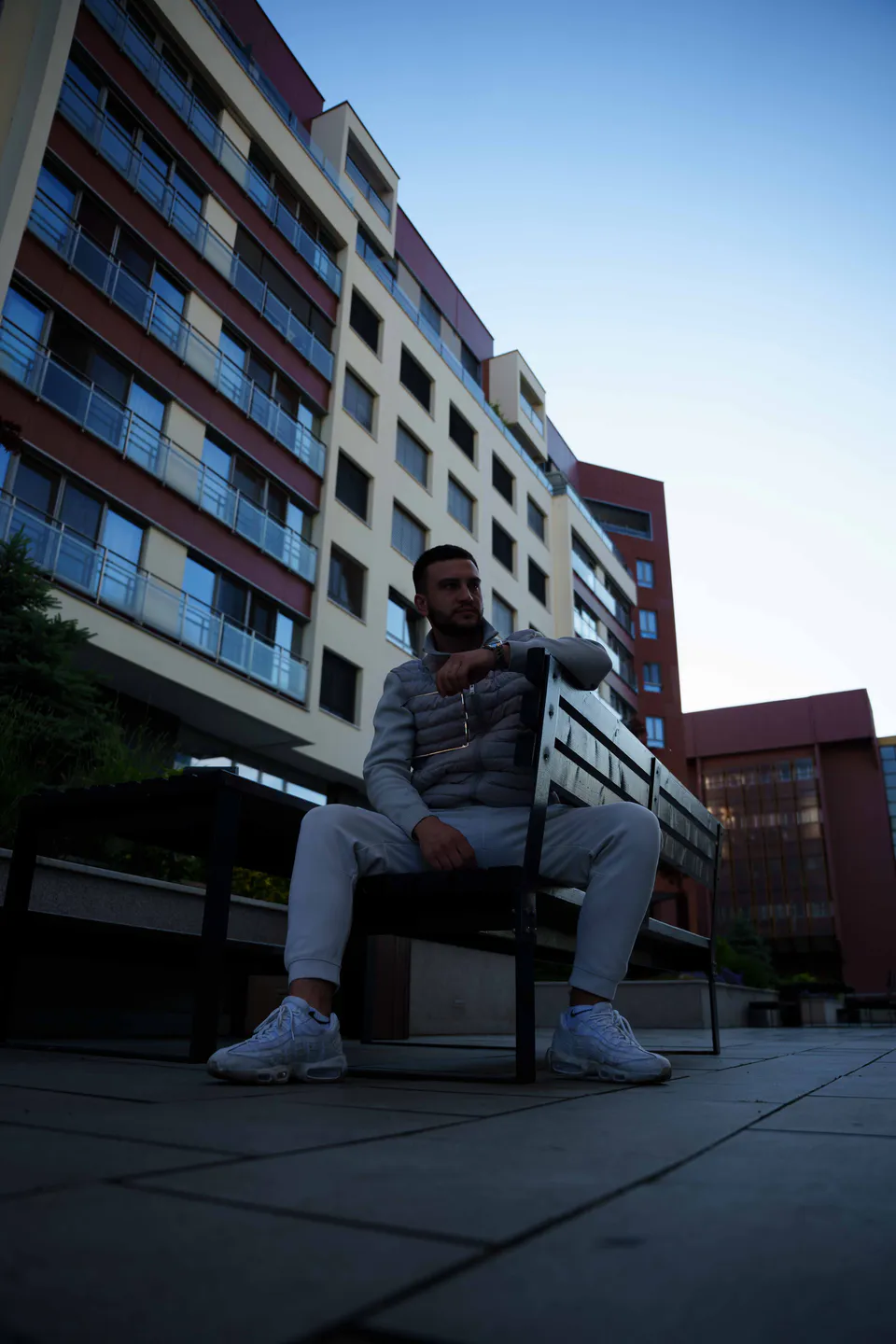 The same man seated on a bench with one arm over the backrest, the office blocks and the last of the dusk sky behind