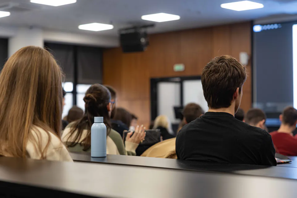Over the shoulders of a lecture-hall audience, a steel water bottle on the desk edge in focus and the room falling away behind it