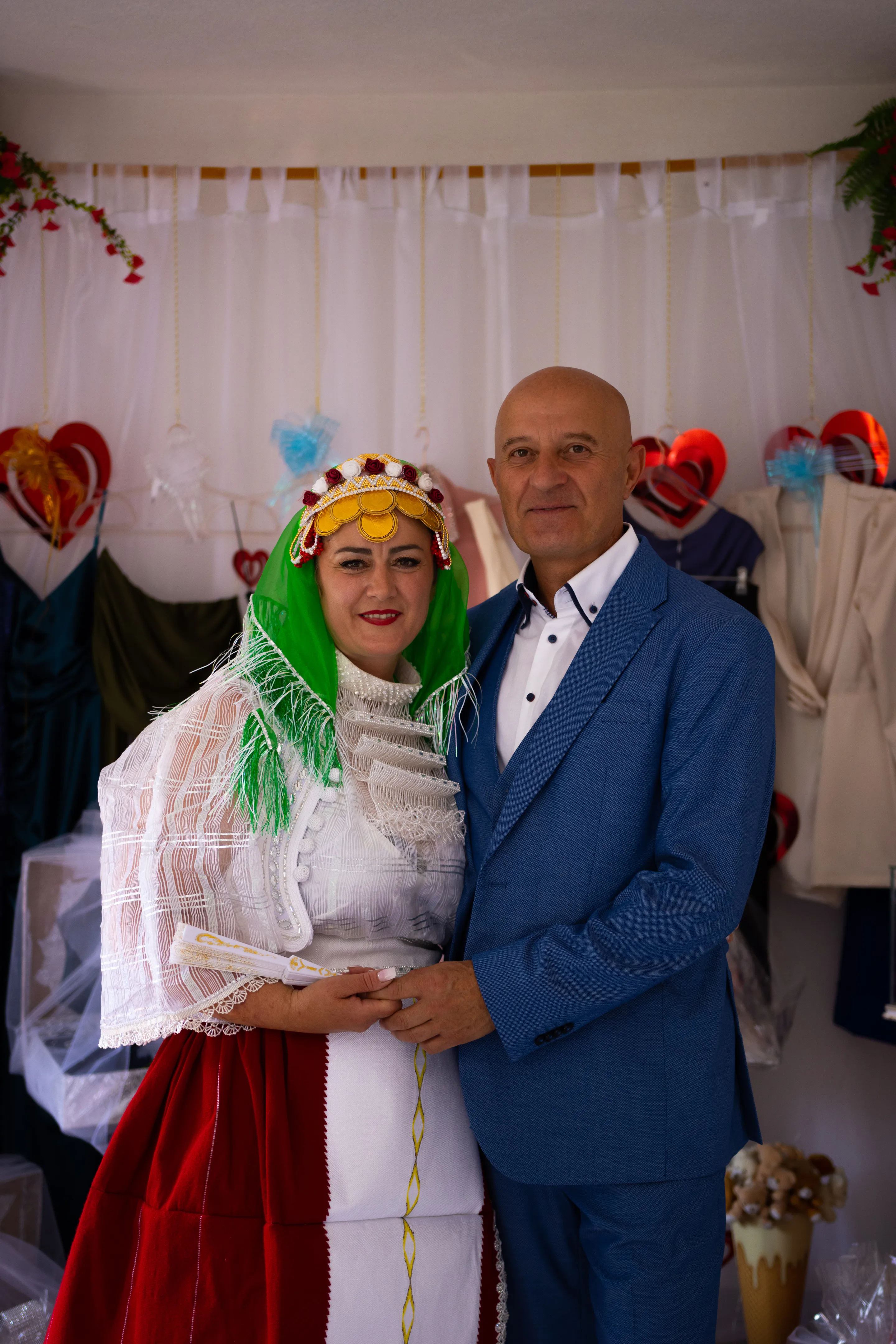 Woman in a gold coin headdress and green scarf beside a man in a blue suit, posed beneath the window
