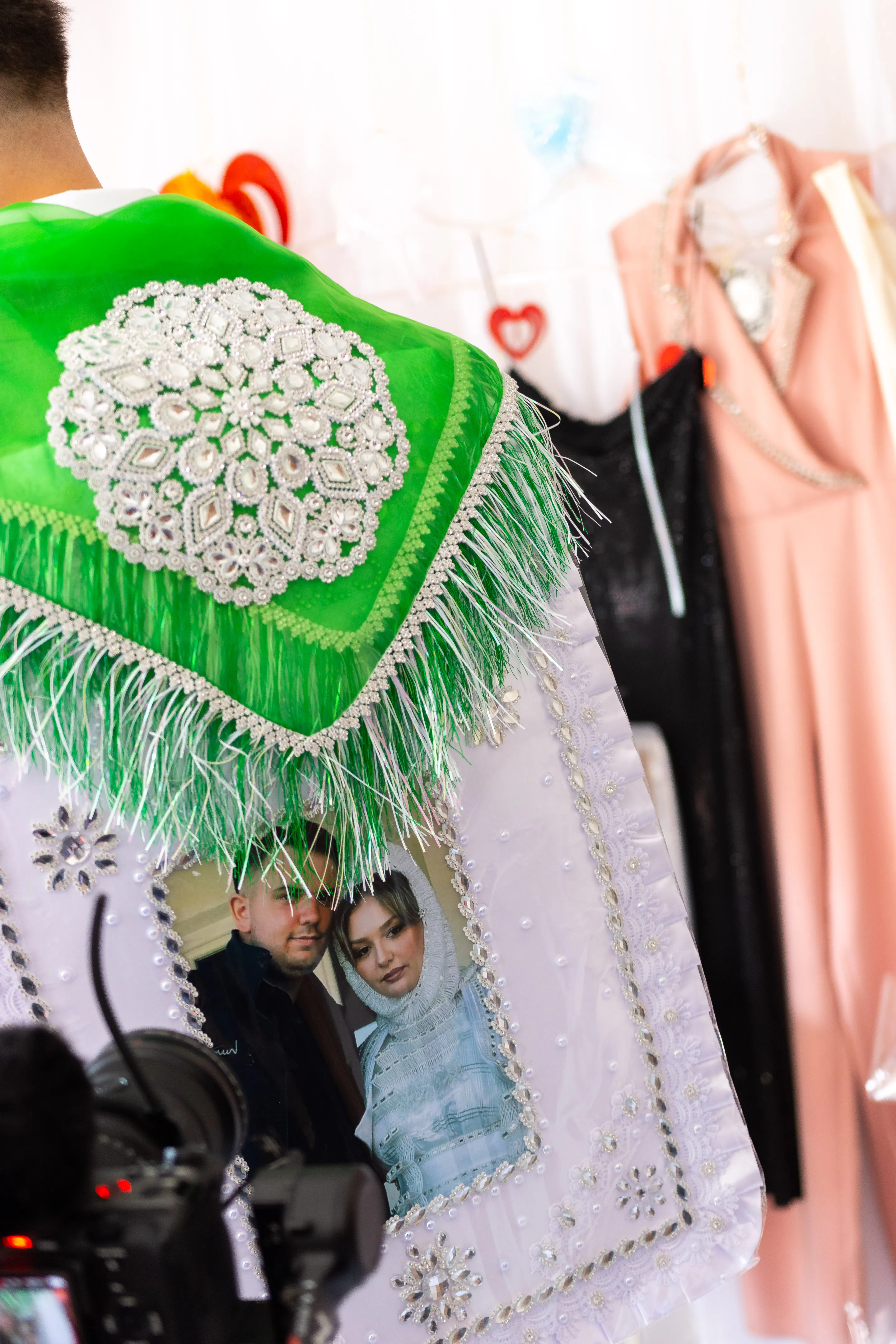 The green fringed shawl with its white beaded medallion hung beside dresses, an embroidered portrait of the couple below