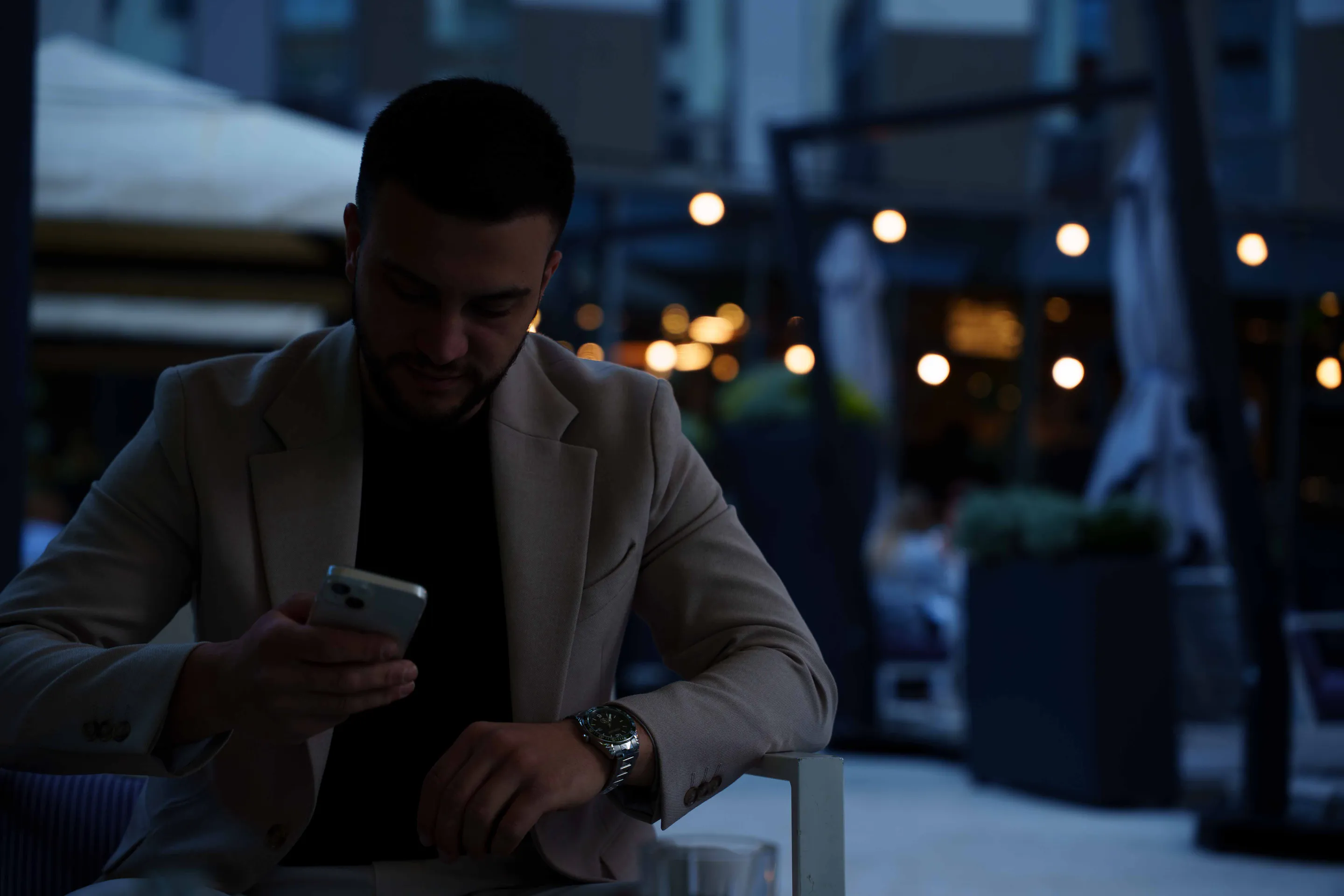 Checking his phone in the dark of the terrace, restaurant lights behind