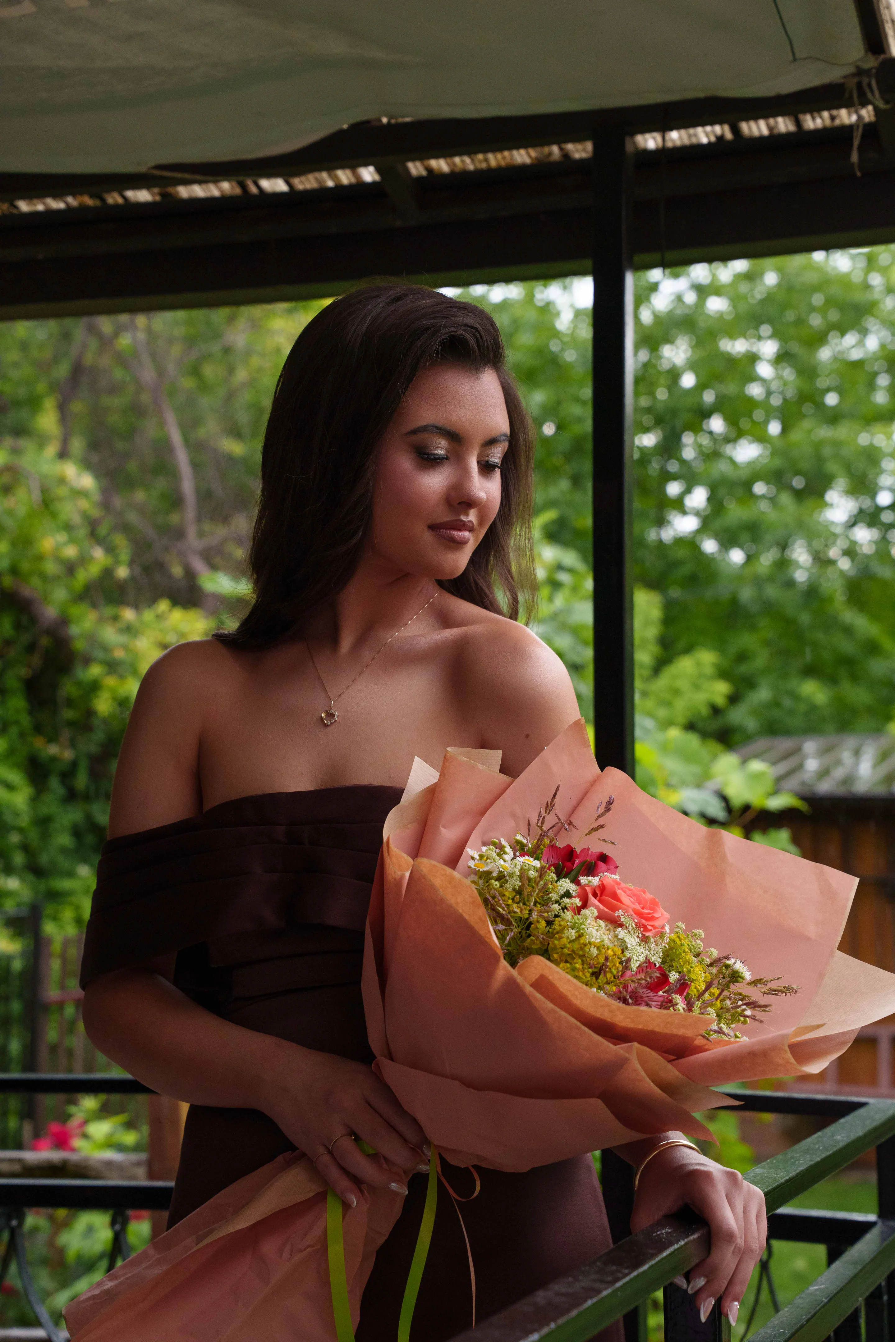 Graduate in a brown satin off-shoulder dress looking down at her bouquet under the pergola