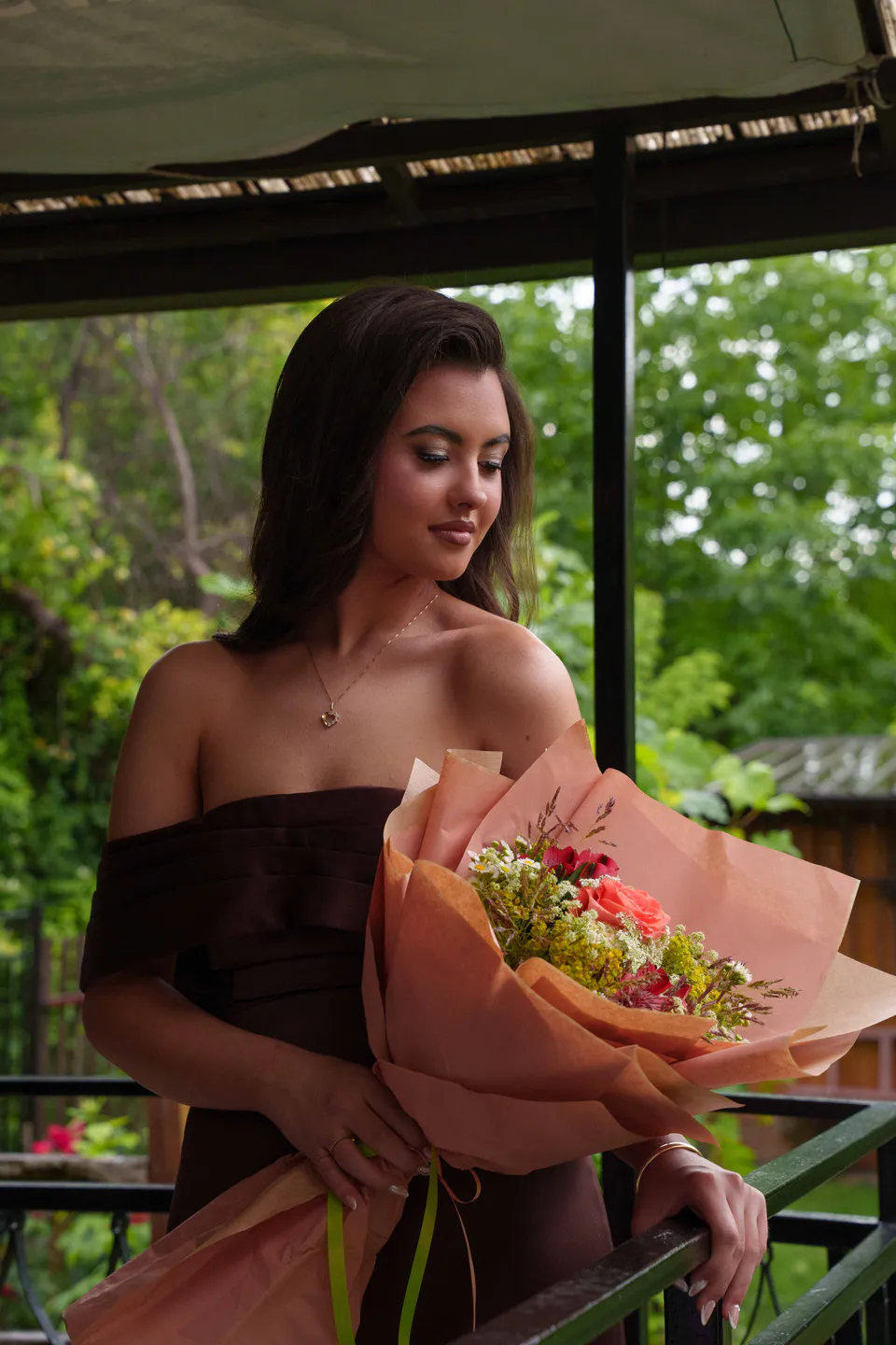 Graduate in a brown satin off-shoulder dress looking down at her bouquet under the pergola