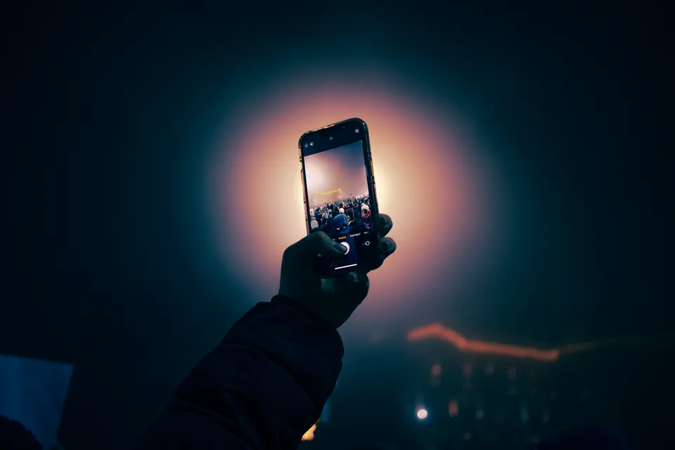 A hand holds a phone up over the crowd, its screen lit against the orange glow
