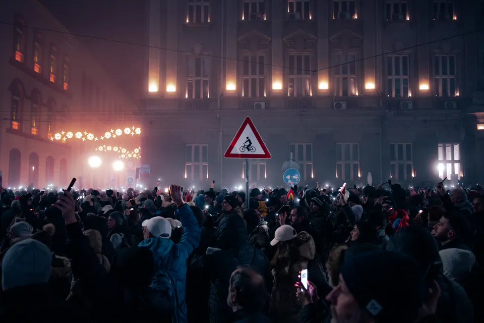 The crowd beneath a lit bicycle-crossing sign, amber windows burning through the fog