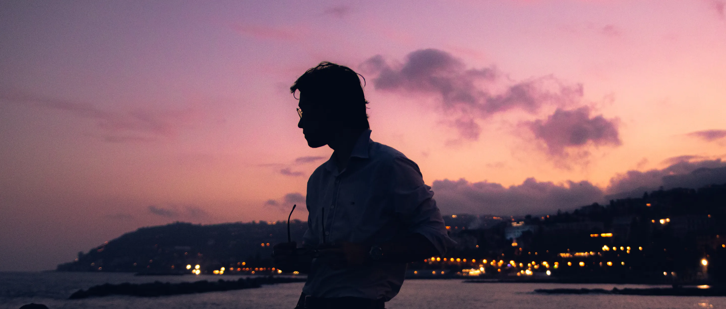 Profile silhouette at dusk, sunglasses in hand, the lit town below