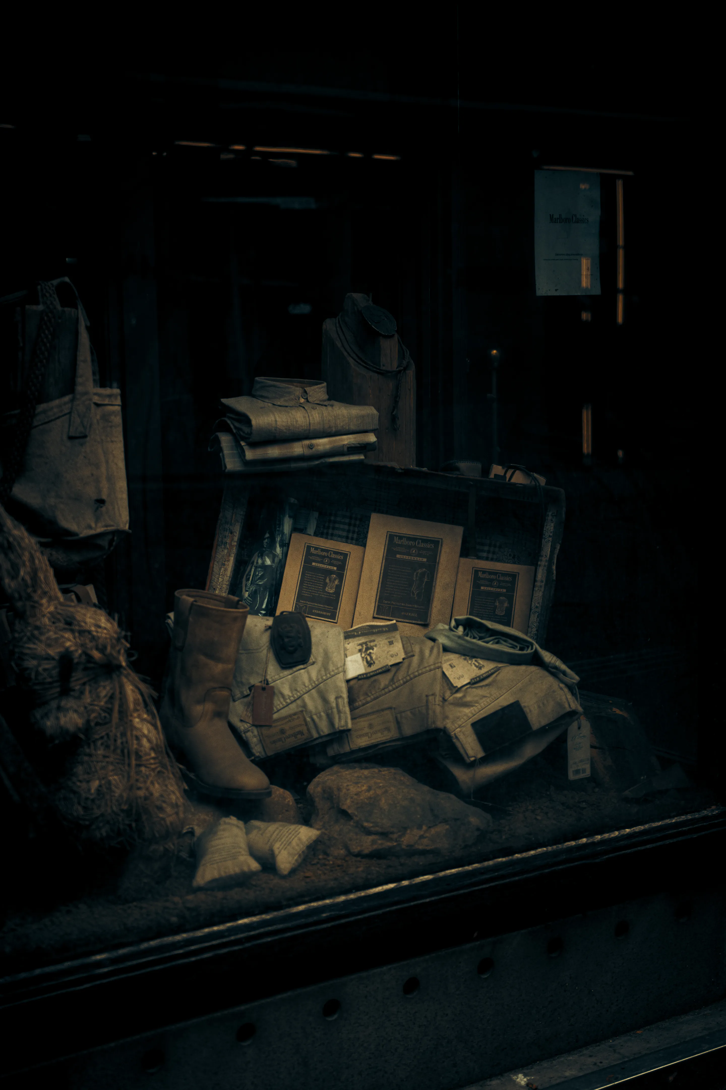 Dim shop window — boots, folded shirts, rope and burlap under a single bulb