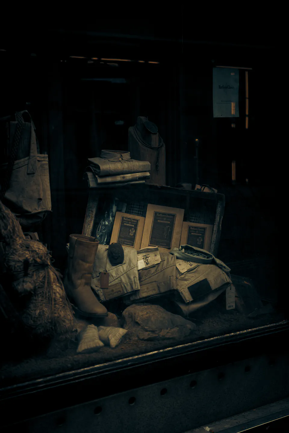 Dim shop window — boots, folded shirts, rope and burlap under a single bulb