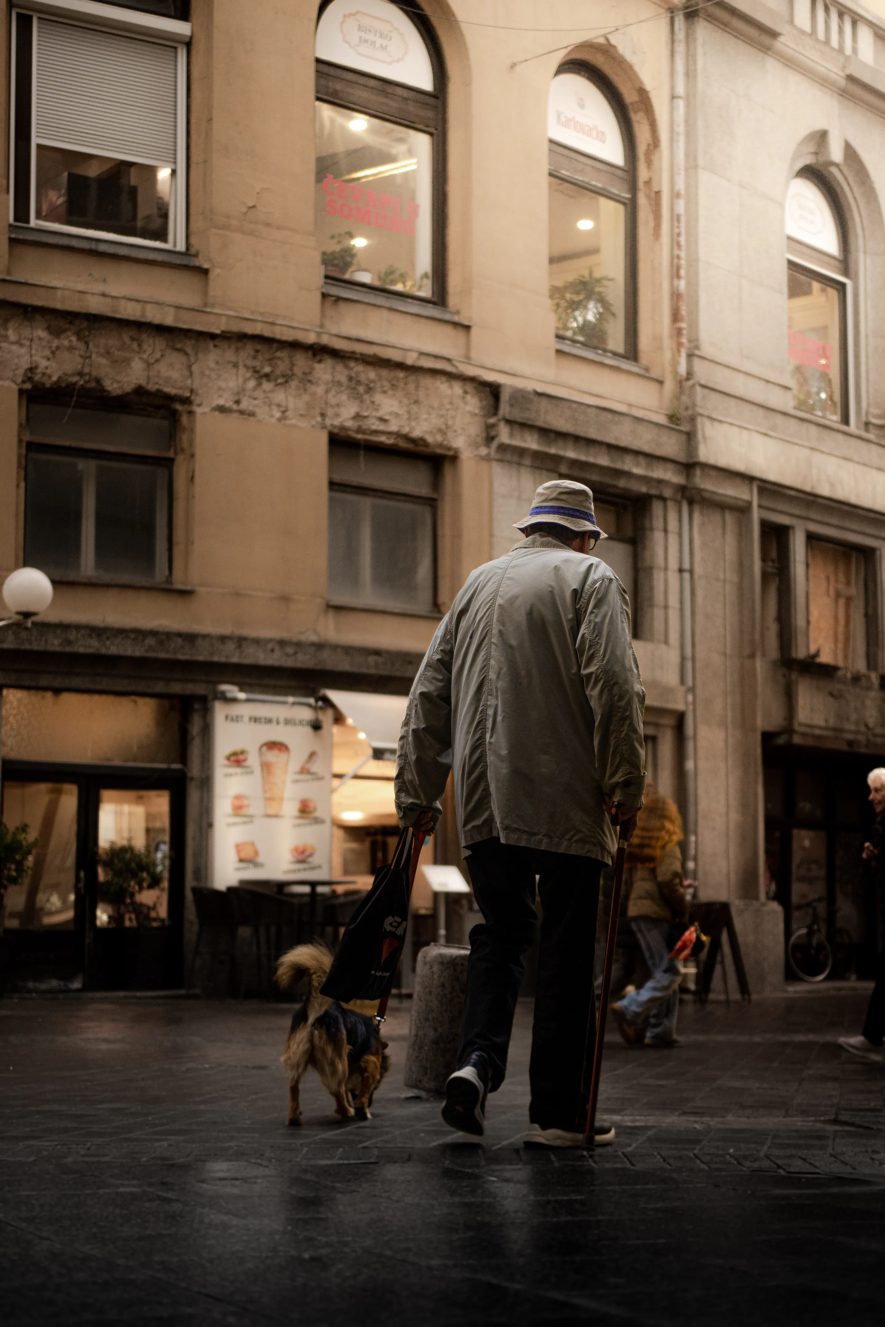 An elderly man in a bucket hat walking his small dog past arched shop windows, cobbles wet