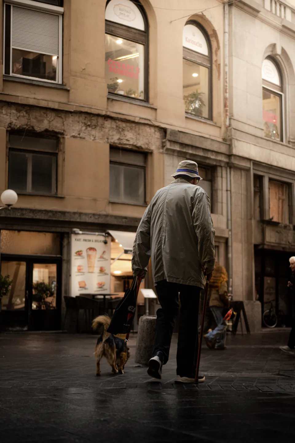 An elderly man in a bucket hat walking his small dog past arched shop windows, cobbles wet