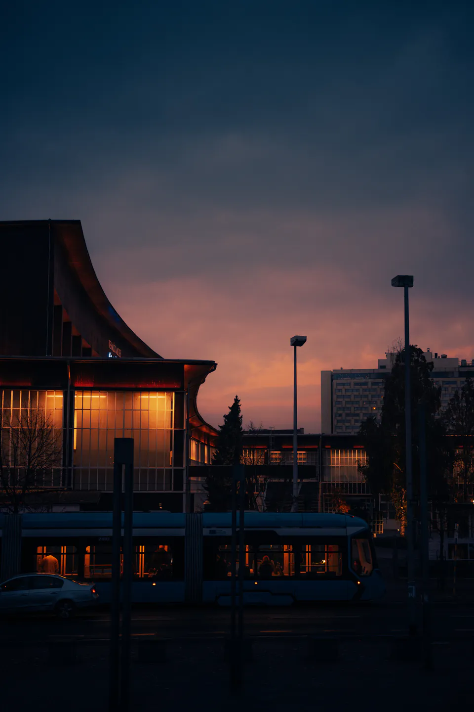 Curved modernist block and a passing tram silhouetted against the orange-to-navy sunset